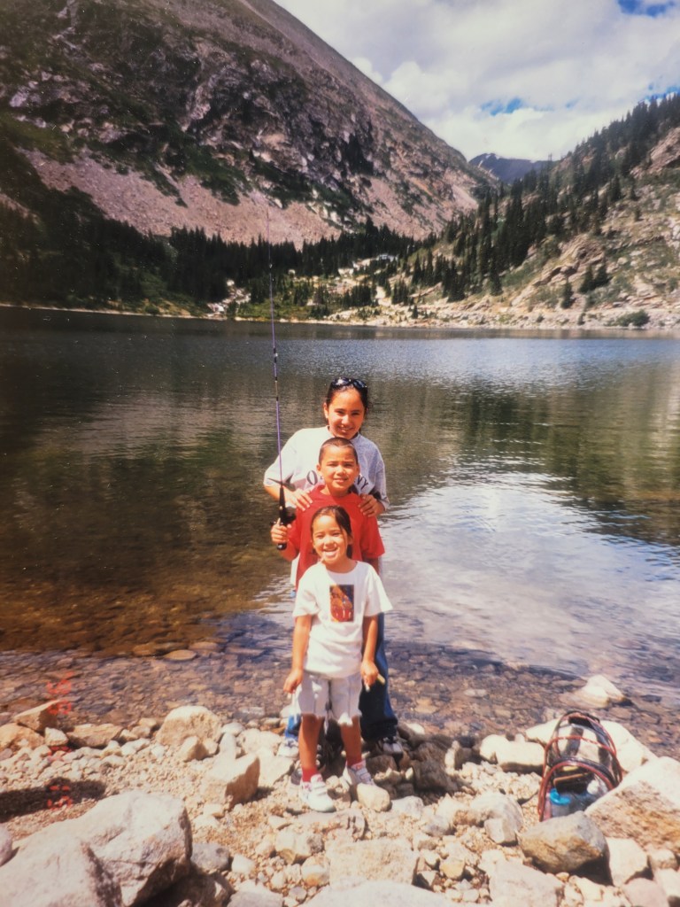 Maya Mineoi as a child with two older siblings stand one in front of each other on rocks at the edge of a lake by the mountains.