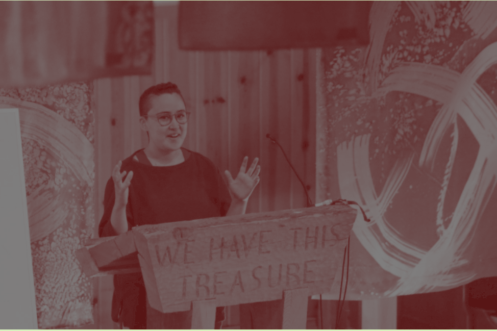 person stands speaking in front of a carved podium which reads, "we have this treasure"