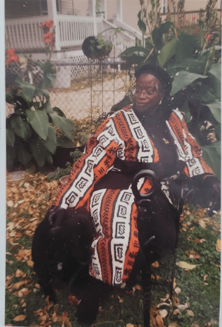 woman sits with a dog in front of large leafed plants. black carved cane in the foreground.